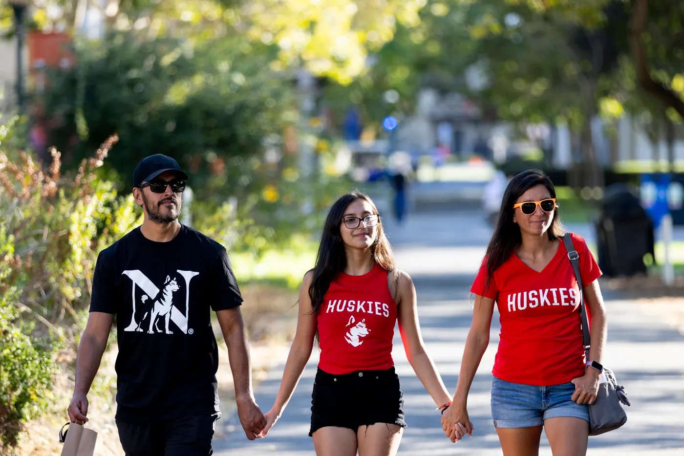 Three people walk along a sunlit, tree-lined campus path, wearing Northeastern branded t-shirts. Academic buildings and other pedestrians are blurred, but visible in the background.